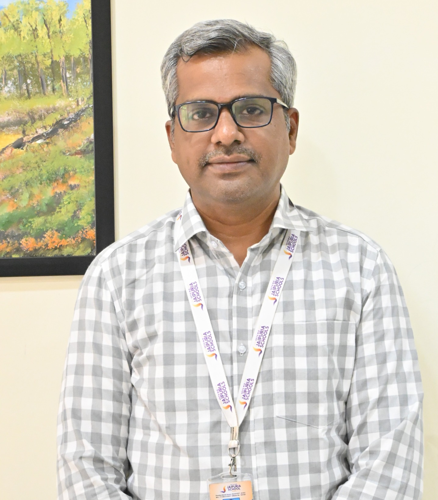 Portrait of a man with gray hair and glasses, wearing a light-gray plaid shirt and a lanyard with Jaipur Schools text, standing indoors near a painting.